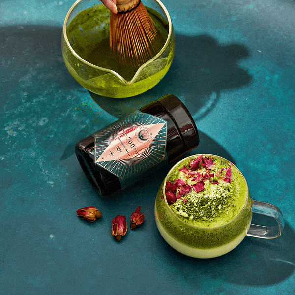 A top-down view of a matcha tea setup on a teal surface. It features a jar of Organic Ceremonial Matcha 700, a matcha whisk in a bowl of green tea, and a cup of Club Magic Hour Tea topped with rose petals and matcha powder. Loose rose petals are scattered around, adding an enchanting touch.