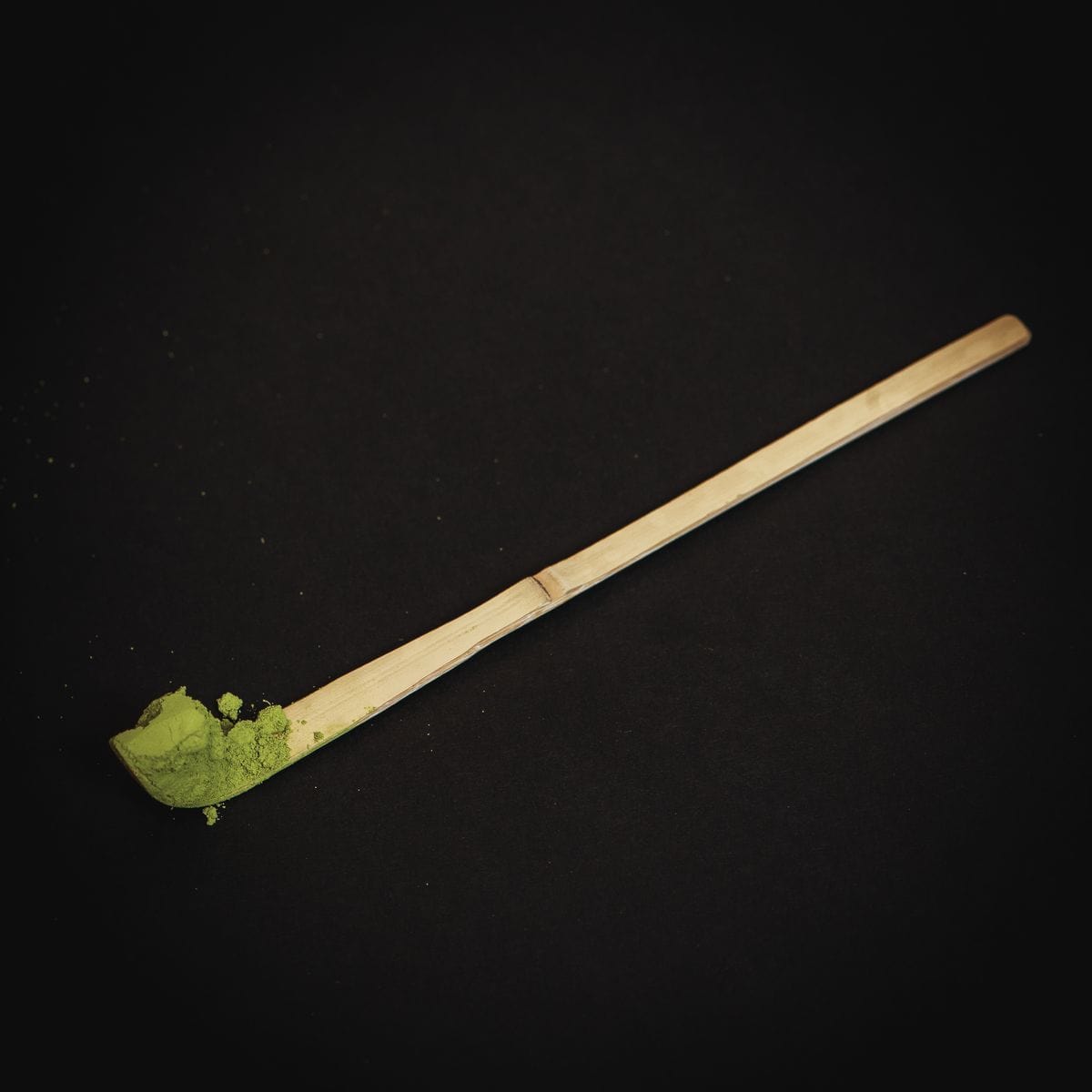 A Ceremonial Matcha Bamboo Scoop by Magic Hour sits on a dark background with some matcha powder on the scoop's end. The green of the matcha contrasts sharply with the dark setting, emphasizing its vibrant color—perfect for enjoying your Magic Hour moment.