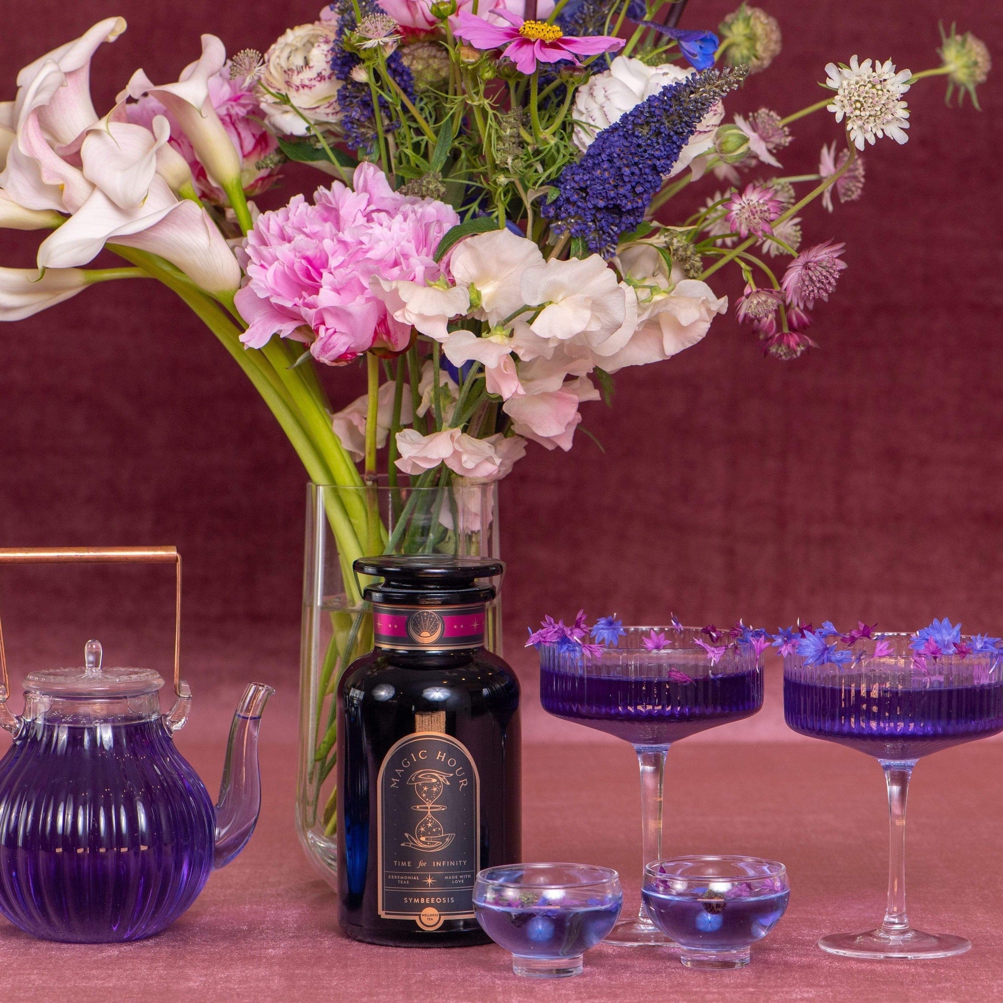 A vibrant setup featuring a dark bottle labeled "Symbeeosis: Beautifying Immunitea for the Queen Bee" from Magic Hour, a purple teapot of loose leaf tea, two purple-tinted glasses with a floral garnish, and a vase of diverse flowers including white lilies and pink peonies, arranged against a rich, burgundy background.