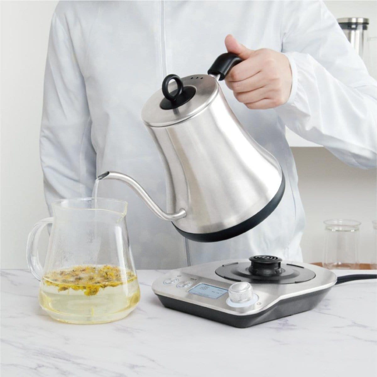 A person in a white jacket is pouring hot water from a Magic Hour Electric Pour Over Kettle into a glass pitcher containing Magic Hour Tea's yellow loose-leaf tea. The kettle, heated on an electric base with digital controls, rests on a white marble countertop.