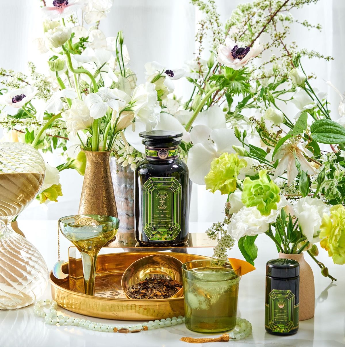 Experience a luxurious tea setup with the Summer Gemstone Wellness Journey - 3-Month Tea Ceremony Box for July, August, and September, featuring a jar adorned with an ornate green label filled with loose leaf tea. This elegant arrangement is complemented by sophisticated glassware and placed on an exquisite gold tray. White and green flowers in vases add a refreshing touch to the scene, enhanced by the presence of a golden hourglass against a well-lit background.