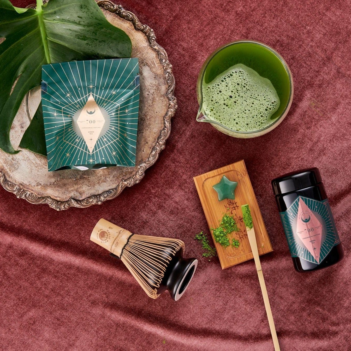 A flat lay image of a traditional matcha tea set, featuring the Chawan Matcha Bowl from Magic Hour. It includes a bamboo whisk, whisk holder, wooden tray with a star-shaped item, silver tray, matcha tea container, and green packaged box filled with loose leaf organic tea.