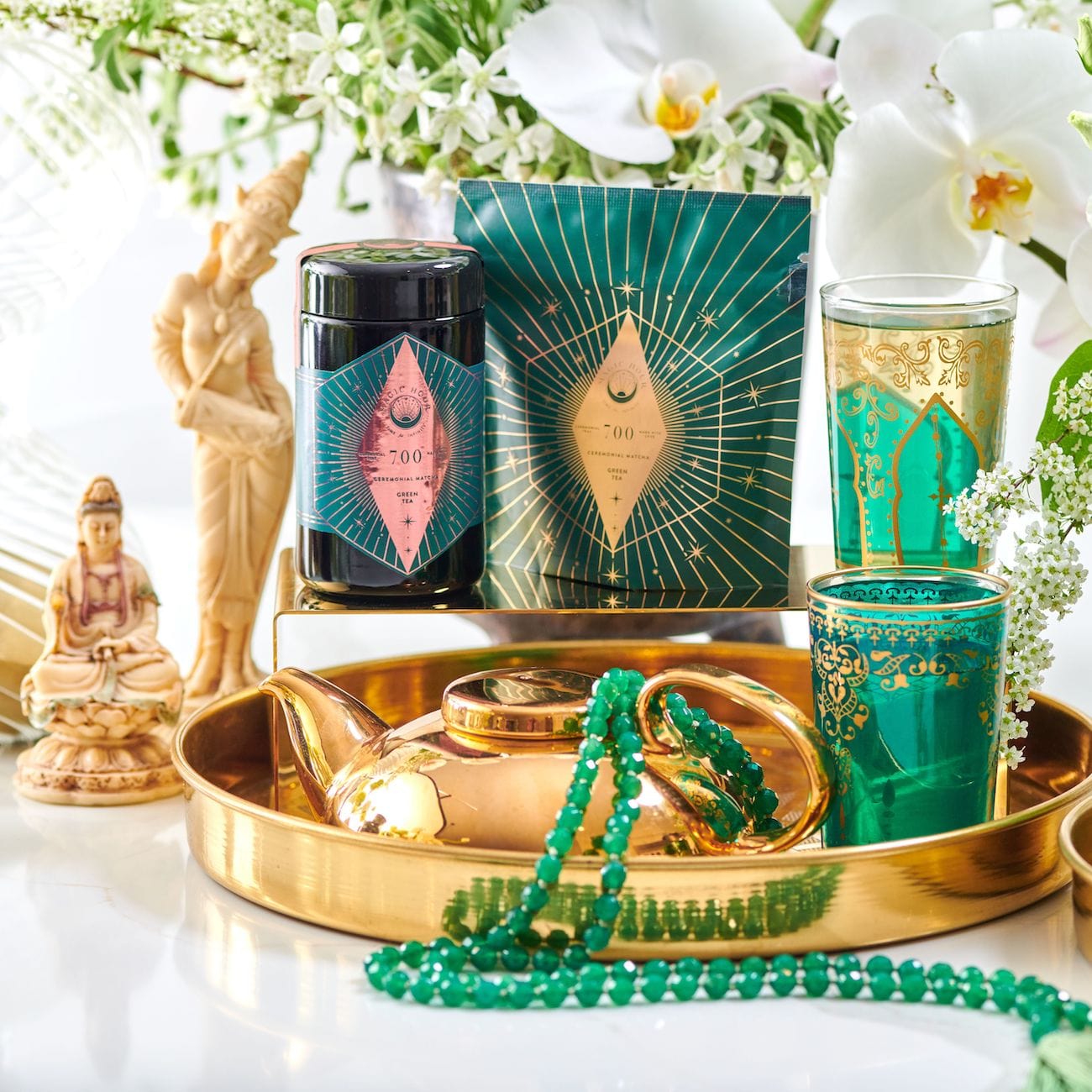 A golden tray displays an assortment of ornate items, including a green and gold teacup, a green beaded necklace, a gold teapot with Organic Ceremonial Matcha 700 from Club Magic Hour, and a jar with a teal and pink label. Floral arrangements with white flowers and statues are in the background.