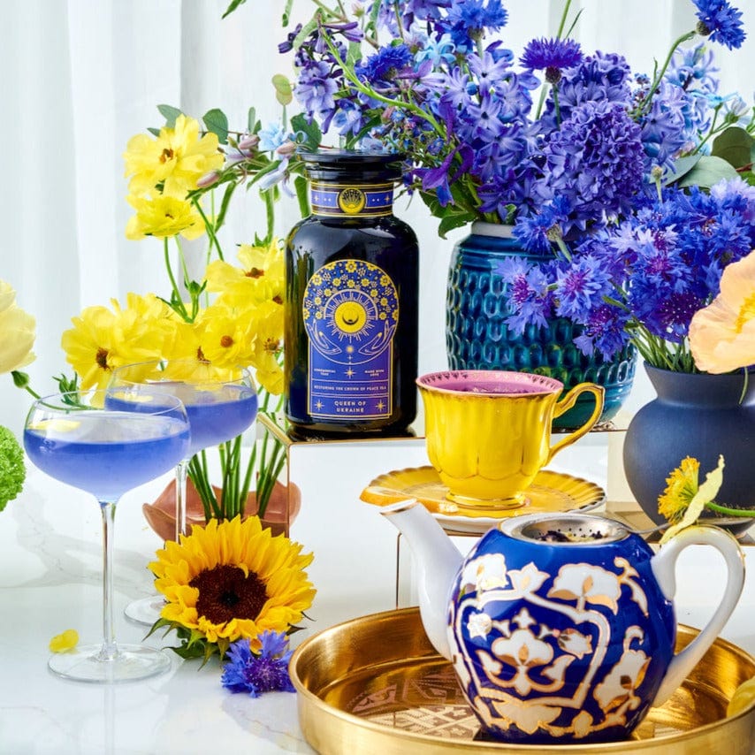 A vibrant arrangement of yellow and purple flowers surrounds an assortment of colorful teacups, a teapot filled with Queen of Ukraine: Spring Blossom Tea by Magic Hour, and a bottle with a decorative label featuring blue and gold designs. A sunflower and a blue cocktail are also present, all set on a reflective white surface.