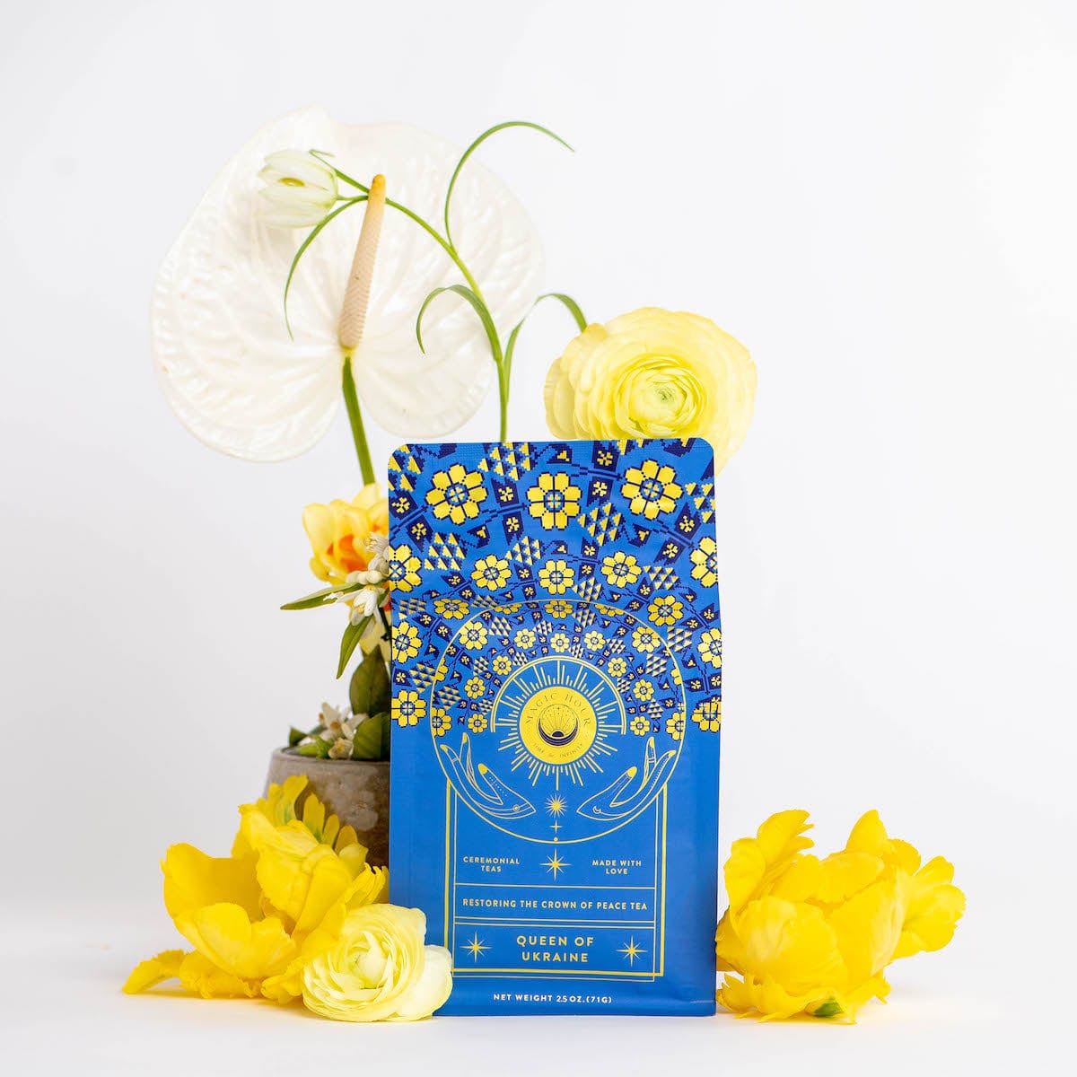 A vibrant blue packet labeled "Magic Hour: Queen of Ukraine: Spring Blossom Tea" with yellow and white floral designs stands against a white background. It is adorned with yellow and white flowers, including roses and anthuriums, enhancing the colorful and elegant presentation of this exquisite loose leaf tea.