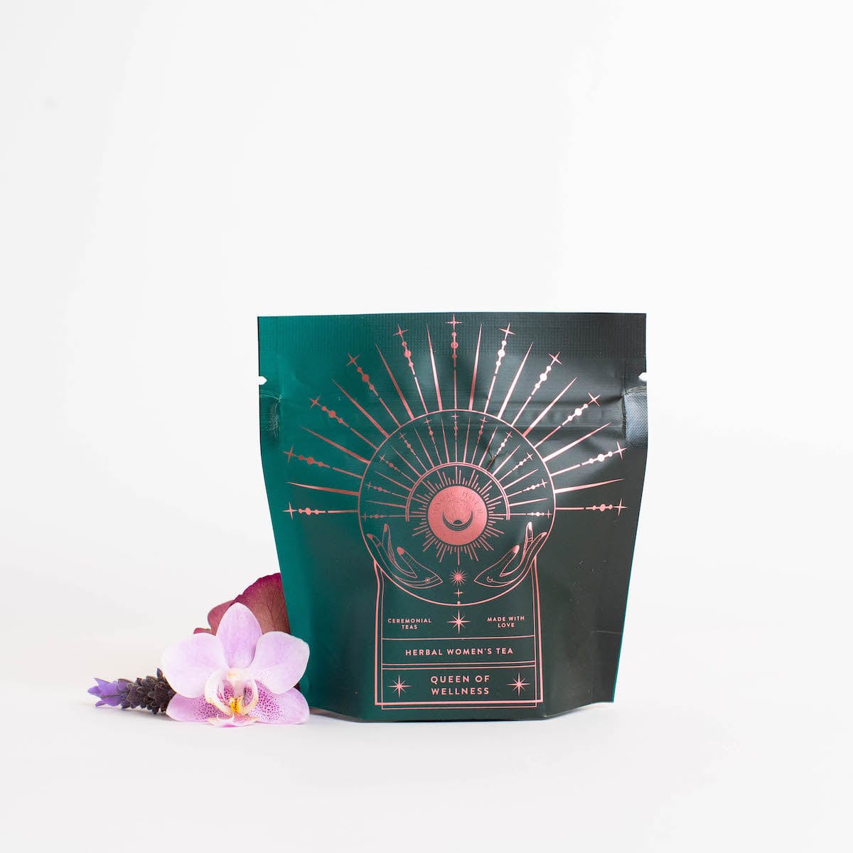 A teal and black package with a celestial design, labeled 'Queen of Wellness: Women's Hormone Balancing Tea for PMS, Healthy Cycles & Menopause,' offers an enchanting experience. Adorned with a golden sunburst and crescent moon, this Magic Hour tea is surrounded by an orchid bloom and lavender sprigs on a white background.