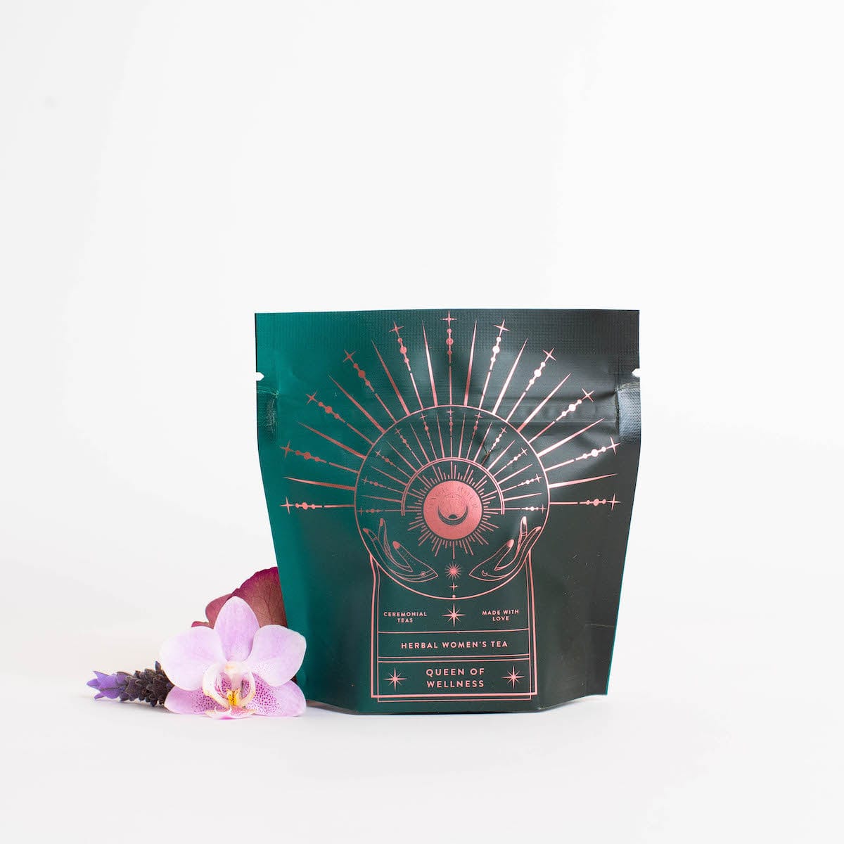 A teal and black package with a celestial design, labeled &quot;Queen of Wellness: Women&#39;s Hormone Balancing Tea for PMS, Healthy Cycles &amp; Menopause,&quot; offers an enchanting experience. Adorned with a golden sunburst and crescent moon, this Magic Hour tea is surrounded by an orchid bloom and lavender sprigs on a white background.