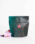 A teal and black package with a celestial design, labeled "Queen of Wellness: Women's Hormone Balancing Tea for PMS, Healthy Cycles & Menopause," offers an enchanting experience. Adorned with a golden sunburst and crescent moon, this Magic Hour tea is surrounded by an orchid bloom and lavender sprigs on a white background.