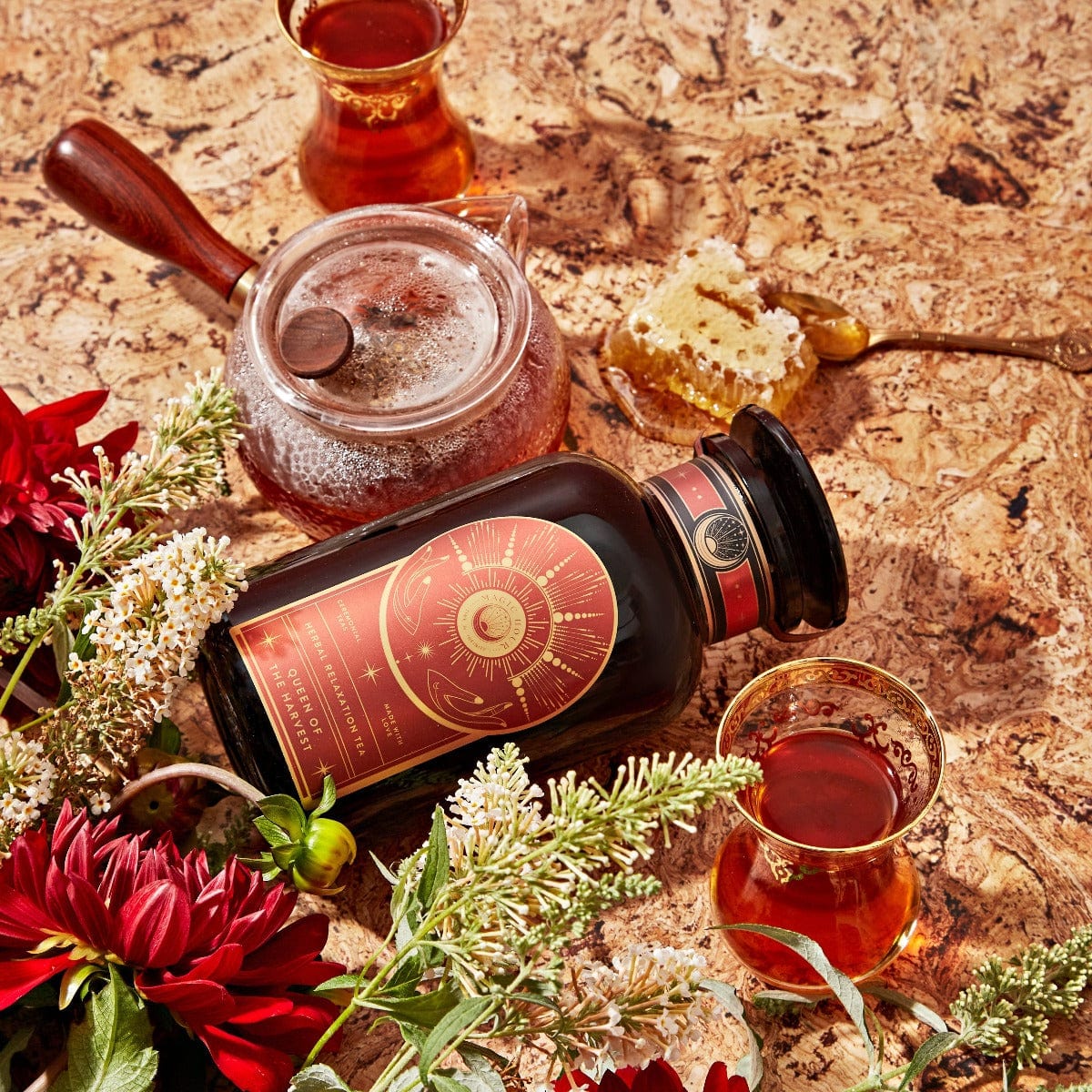 A box of Magic Hour&#39;s Queen of the Harvest: Herbal Relaxation Tea is surrounded by a glass teapot filled with organic tea, a honeycomb with a golden spoon, two traditional tea glasses brimming with loose leaf tea, red and white flowers, and greenery on a speckled countertop. The scene exudes a cozy and inviting atmosphere.