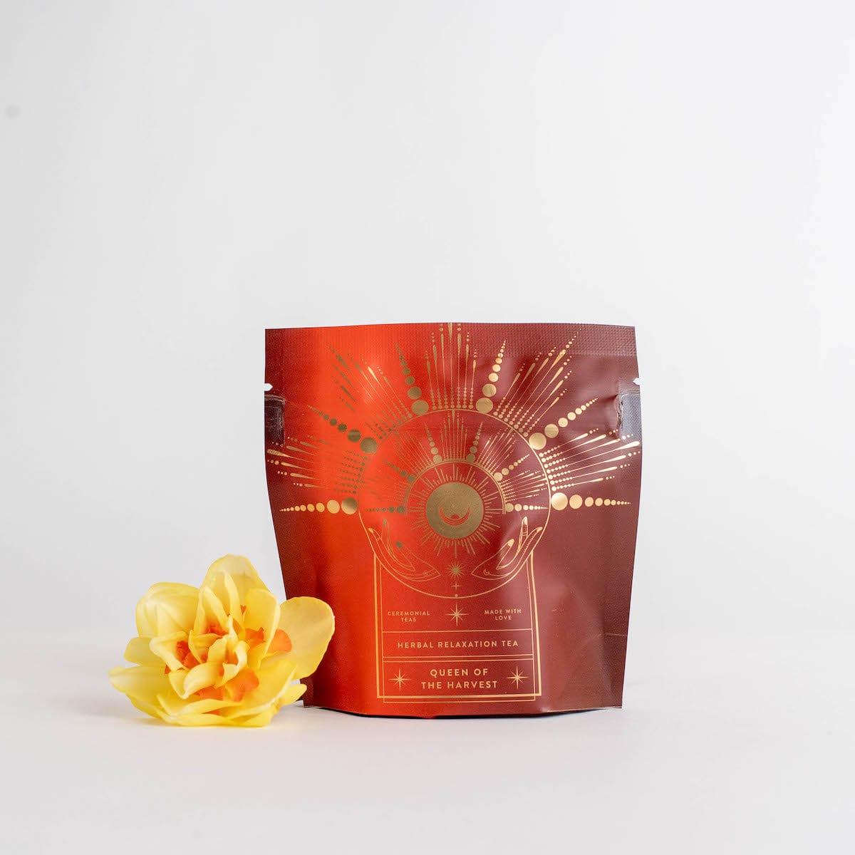 A pouch of **Queen of the Harvest : Herbal Relaxation Tea** labeled **Magic Hour** with an intricate gold design stands against a white background. To the left of the pouch, there is a vibrant yellow flower placed for decoration, enhancing the allure of this organic loose leaf tea.