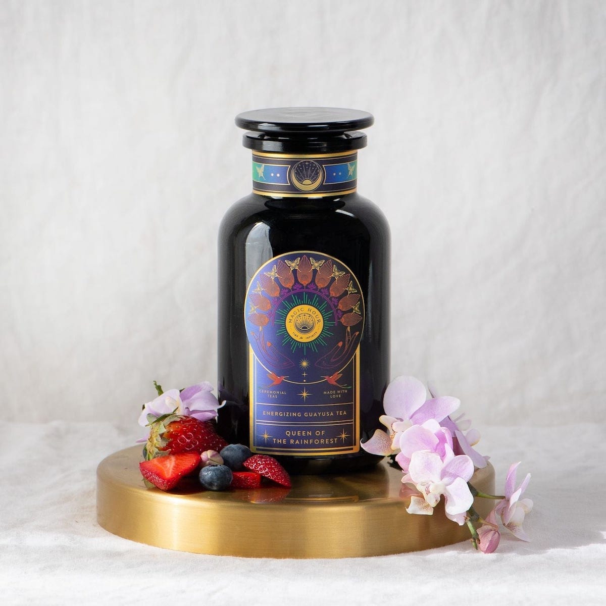 A dark glass jar labeled "Queen of the Rainforest : Cacao-Berry Tea" by Magic Hour sits on a circular wooden stand, surrounded by fresh strawberries, blueberries, and delicate purple and white flowers. The organic tea promises a delightful experience. In the background is a soft white cloth, enhancing the serene ambiance.