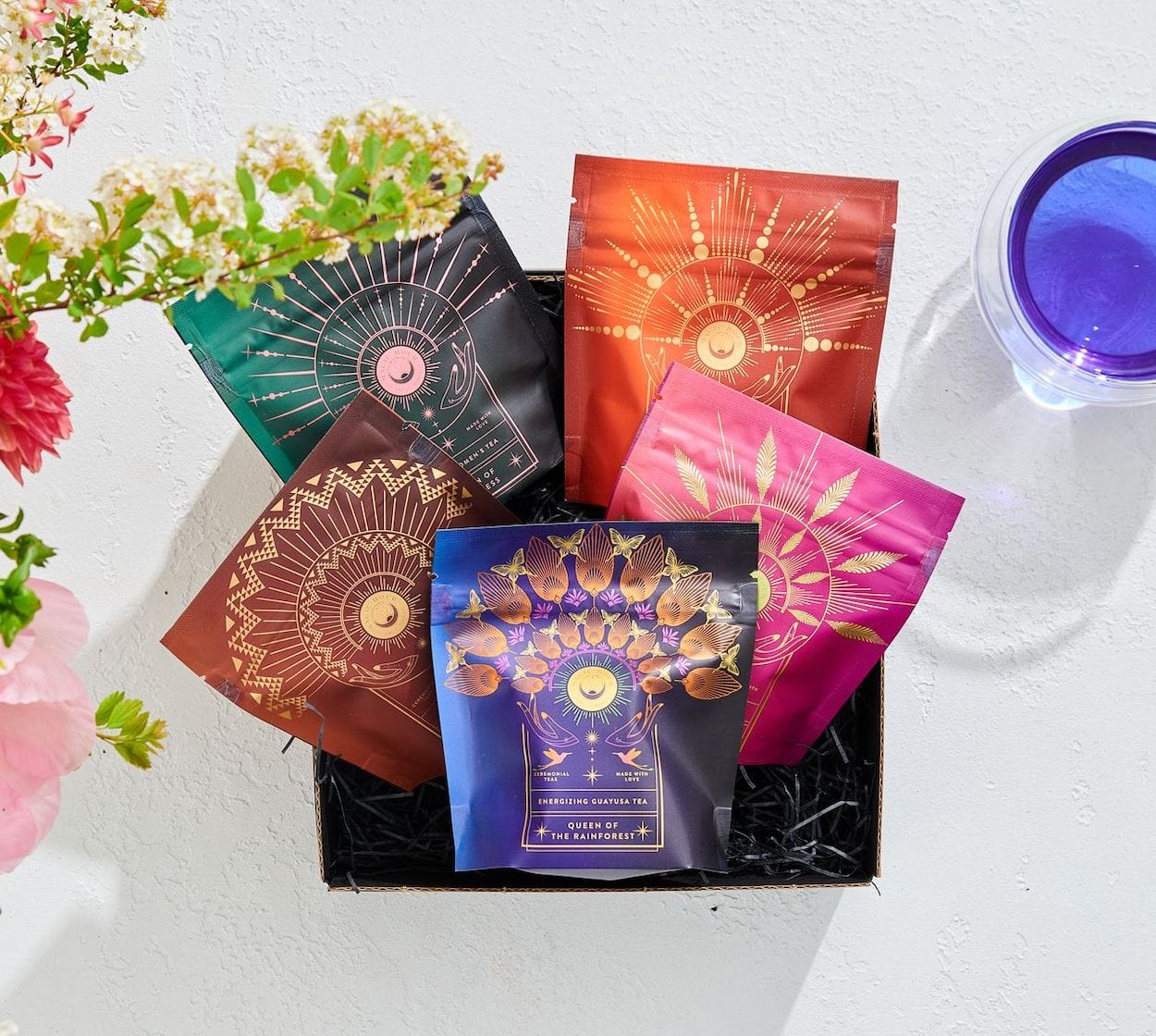 A black gift box containing five colorful packets with intricate gold designs is displayed on a white surface. Surrounding the box are a pink flower and green foliage on the left and a purple-tinted drinking glass on the right, perfect for savoring The Queen's Sampler Set by Magic Hour.