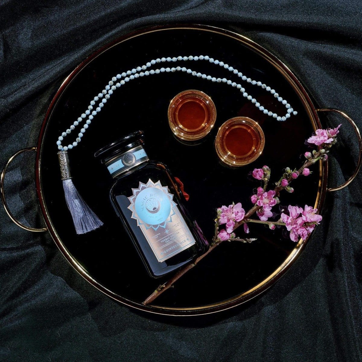 A decorative tray holds a perfume bottle with a light blue tassel, a string of white beads, two small cups filled with Magic Hour's Quintessence™ Tea for Opening & Healing the Throat Chakra amber liquid, and a sprig of pink flowers. The tray, adorned with golden handles, rests elegantly on dark, textured fabric.