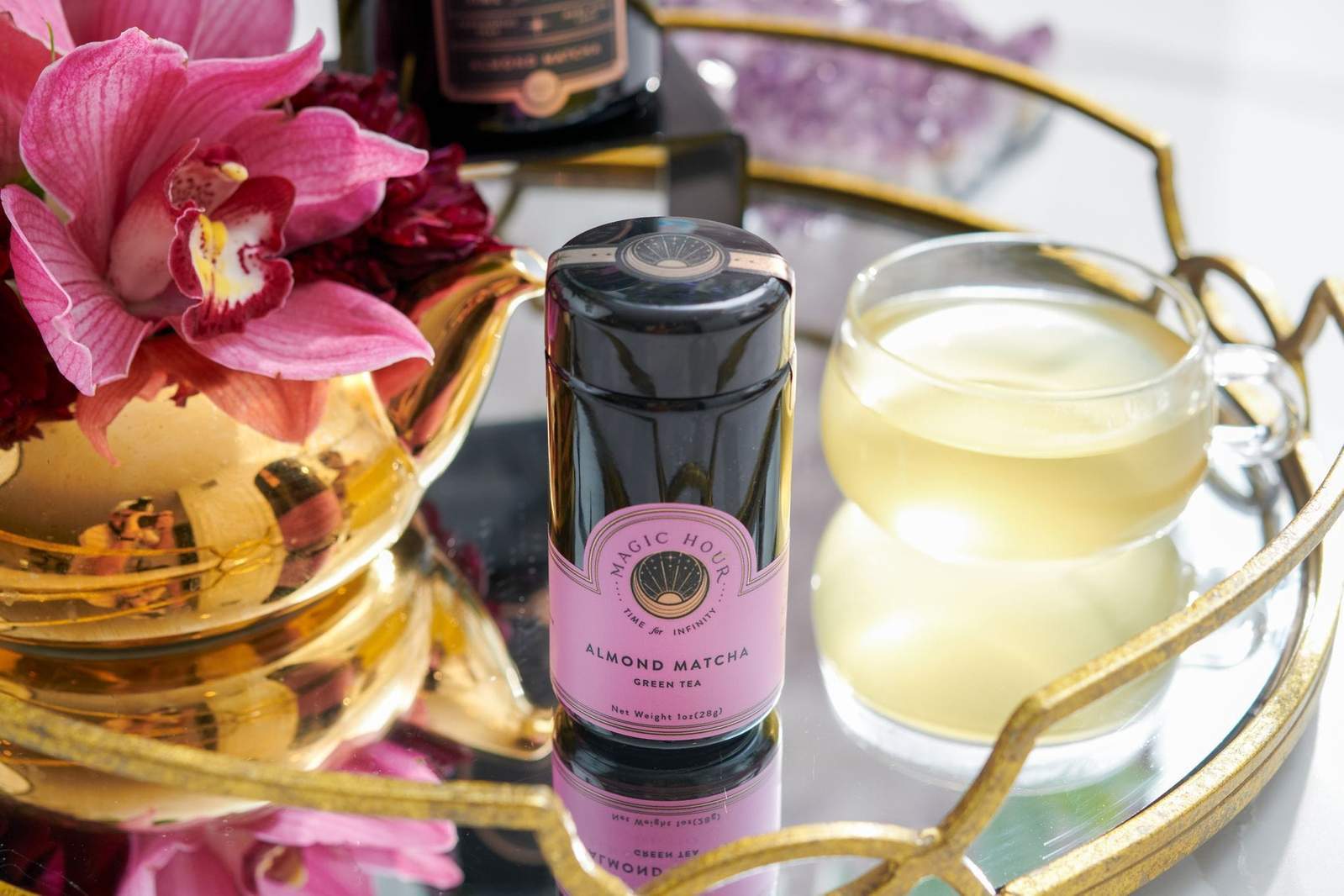 A jar of Magic Hour Sip Wellness: Ritual Kit for Immunity & Joy is placed on a gold-trimmed tray next to a transparent teacup filled with organic tea. Nearby, a pink orchid arrangement and a piece of amethyst decoratively complement the scene.