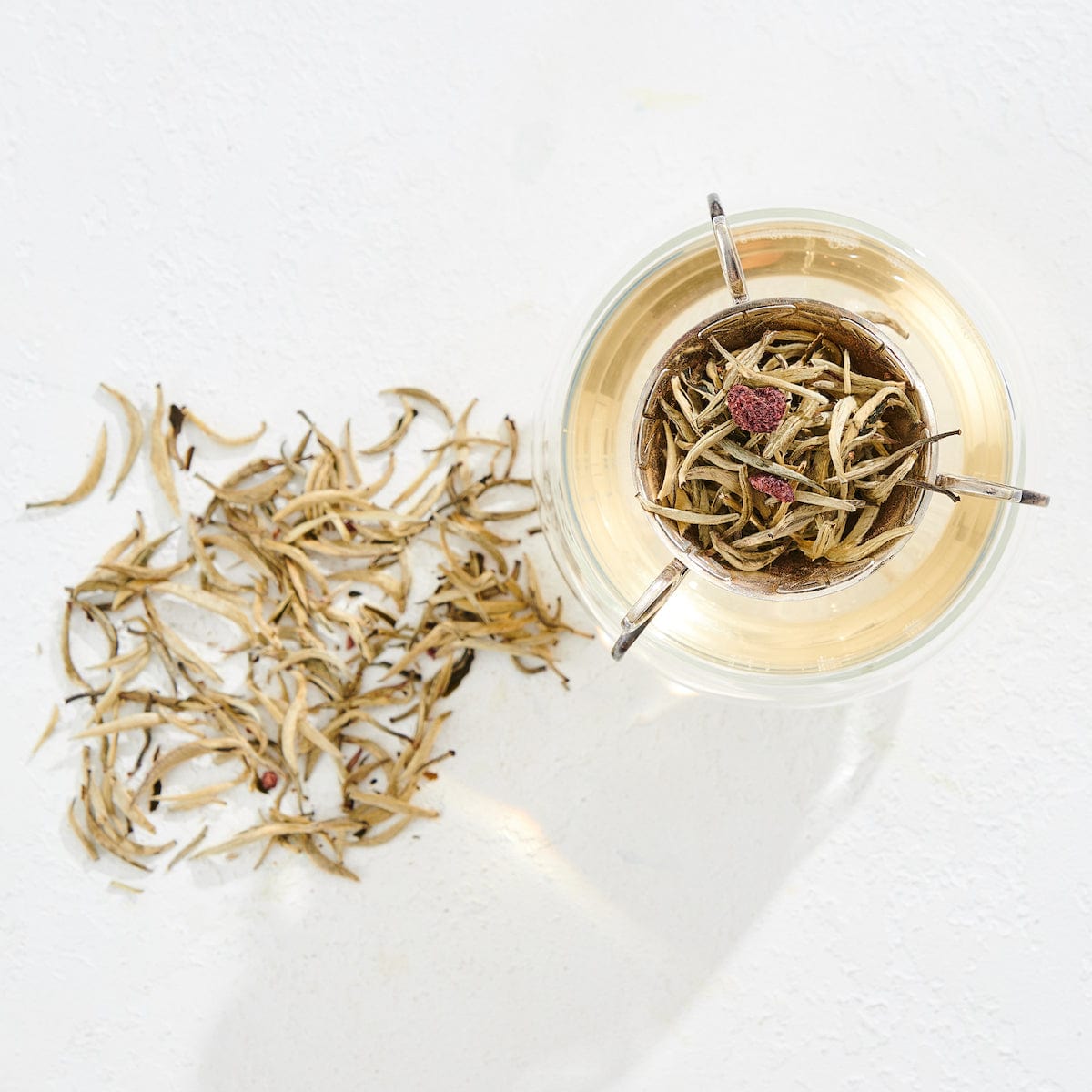 Overhead view of a cup filled with light-colored organic tea, with a metal strainer holding loose tea leaves inside it. To the left of the cup, a small pile of dry tea leaves is scattered on a white surface. The scene is brightly lit, capturing the essence of Club Magic Hour Raspberry Earl White Tea.