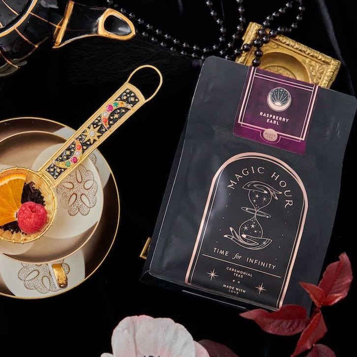 A beautifully arranged flatlay showcases a bag of Raspberry Earl Grey Black Tea from Club Magic Hour, an ornate tea strainer with loose leaf tea and an orange slice, a cup on a saucer, black beads, and a small gold frame against a black fabric backdrop.