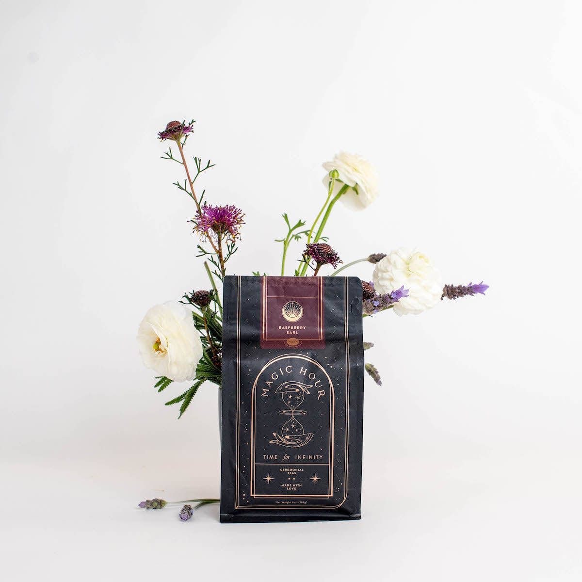 A dark-colored packet labeled "Club Magic Hour: Raspberry Earl White Tea" is placed against a white background, partially surrounded by a small arrangement of white and purple flowers. The packet, filled with organic loose leaf tea, displays an intricate design featuring celestial imagery.