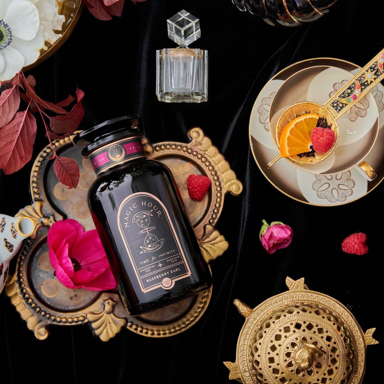 A decorative scene featuring a bottle of Club Magic Hour Raspberry Earl Grey Black Tea on an ornate platter with various decorative items around it, including a teacup with an orange slice, raspberries, a flower, and a golden tea strainer on a black velvet backdrop.