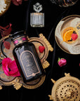 A decorative scene featuring a bottle of Club Magic Hour Raspberry Earl Grey Black Tea on an ornate platter with various decorative items around it, including a teacup with an orange slice, raspberries, a flower, and a golden tea strainer on a black velvet backdrop.