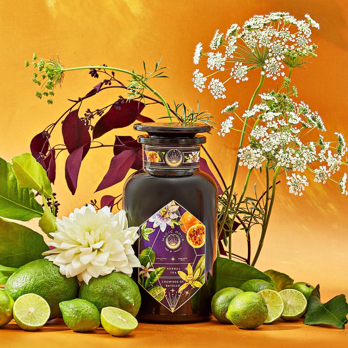 An elegant dark glass bottle labeled "Romance in Ravello" from Magic Hour is surrounded by fresh green limes, lime wedges, and assorted flowers against a vibrant yellow-orange background. This visually rich composition evokes the natural beauty and freshness of the Amalfi Coast, enhanced by hints of chocolate, passion fruit, and rose cream.