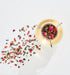 A glass tea infuser filled with loose leaf tea from the Classics - Luxe Sampler Sips collection, showcasing visible flower petals and herbs, sits on a white surface. Some of the colorful tea leaves and petals are scattered around the infuser, creating a vibrant visual contrast with the background.