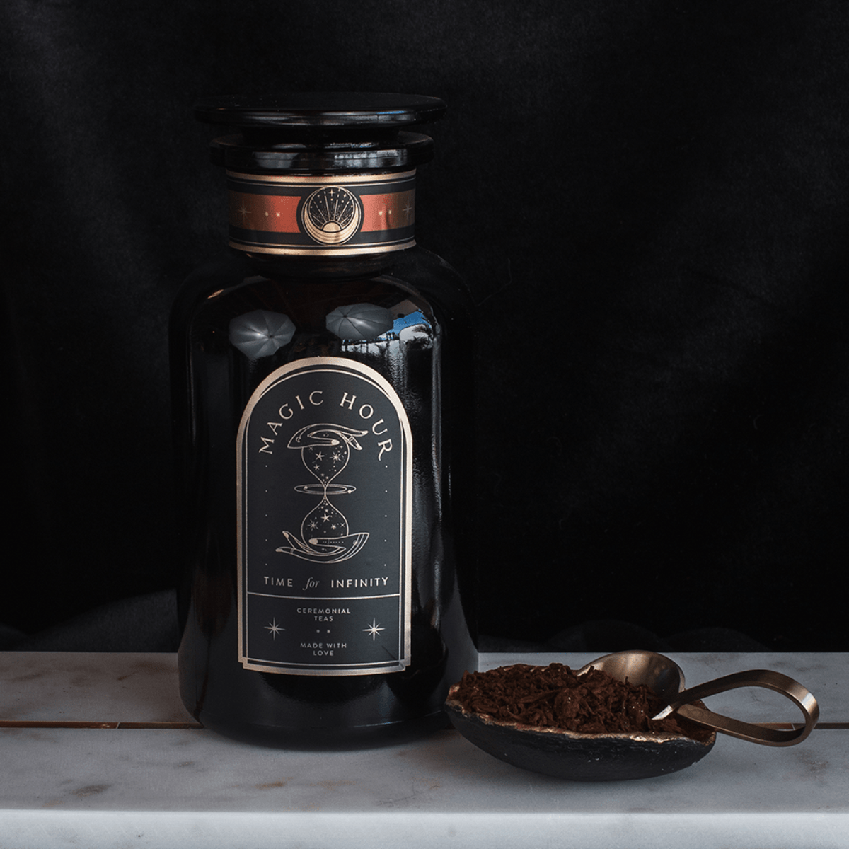 A black glass jar labeled "Club Magic Hour Rise & Shine Black Tea" with an illustration of an hourglass, set against a dark background. Next to the jar is a gold spoon resting in an oval dish filled with loose leaf tea, possibly organic tea.