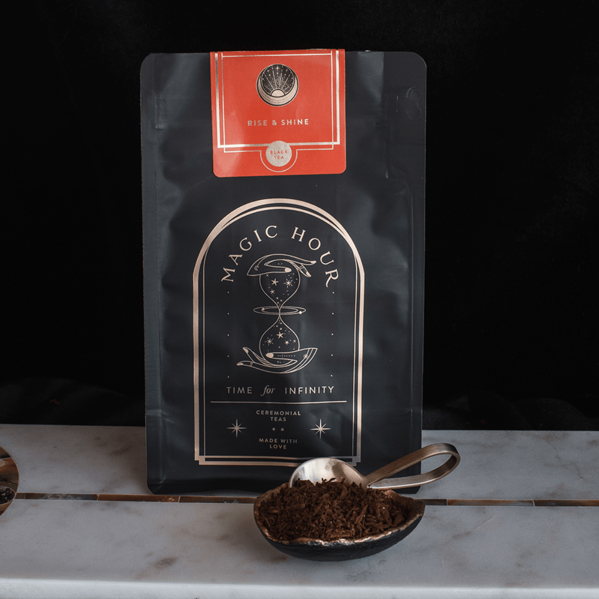 A black, resealable bag labeled "Rise & Shine Black Tea" with a celestial design and the phrase "Time for Infinity" is displayed upright. A red label at the top says "Club Magic Hour." In front, a spoon rests in a small bowl filled with organic loose leaf tea.