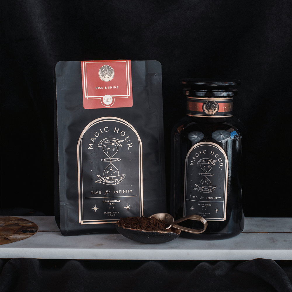 A photo of two Club Magic Hour products. On the left is a flat pouch labeled "Rise & Shine Black Tea," featuring organic loose leaf tea. On the right is a black jar labeled "Time & Infinity," with both items adorned with celestial-themed designs and a teaspoon filled with tea leaves placed in front.
