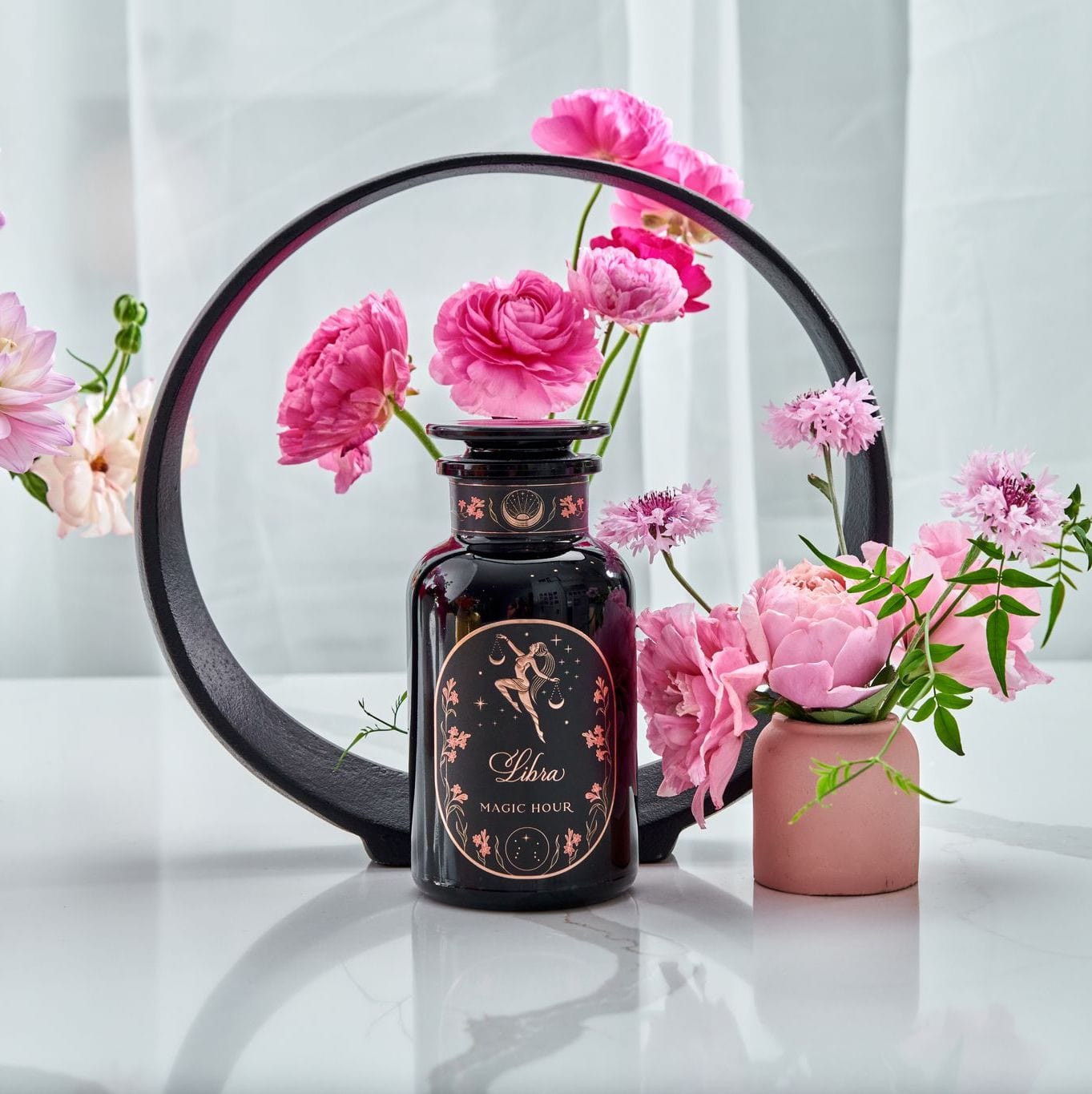 A black ring stand holds pink flowers, framing a dark bottle labeled "Magic Hour Libra- Pistachio-Rose Persian Love Cake Tea with White Pearls & Shatavari" in the foreground. The bottle has a floral design and is accompanied by more pink flowers in a small vase on the right. White fabric drapes in the background, adding an elegant touch to this scene.
