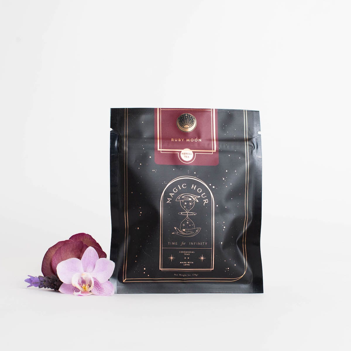 A black and gold pouch labeled &quot;Ruby Moon™ : Hibiscus Elderberry Tea&quot; rests against a white background. The pouch, containing organic loose leaf tea by Club Magic Hour, features galaxy-themed designs. Beside it lies a small arrangement of purple and white orchids.