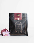 A black and gold pouch labeled "Ruby Moon™ : Hibiscus Elderberry Tea" rests against a white background. The pouch, containing organic loose leaf tea by Club Magic Hour, features galaxy-themed designs. Beside it lies a small arrangement of purple and white orchids.