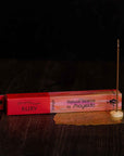 A stick of burning sandalwood incense stands upright in a small holder on a dark wooden surface. Positioned next to it is a red and pink box labeled "Natural Incense : Ruby" by Shoyeido. A decorative golden mesh lies beneath the holder.