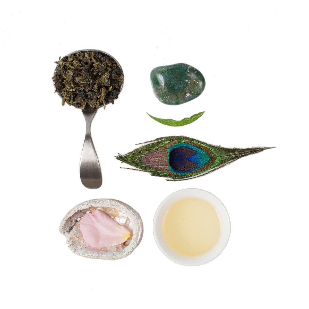 A composition displaying a spoonful of Ti Quan Yin Oolong: Tea of Patient Compassion leaves, a green gemstone, a peacock feather, a fresh leaf, a polished shell with a pink rose petal inside, and a cup of light-colored tea. All arranged neatly on a white background to highlight the wellness benefits of Magic Hour Ti Quan Yin Oolong: Tea of Patient Compassion.
