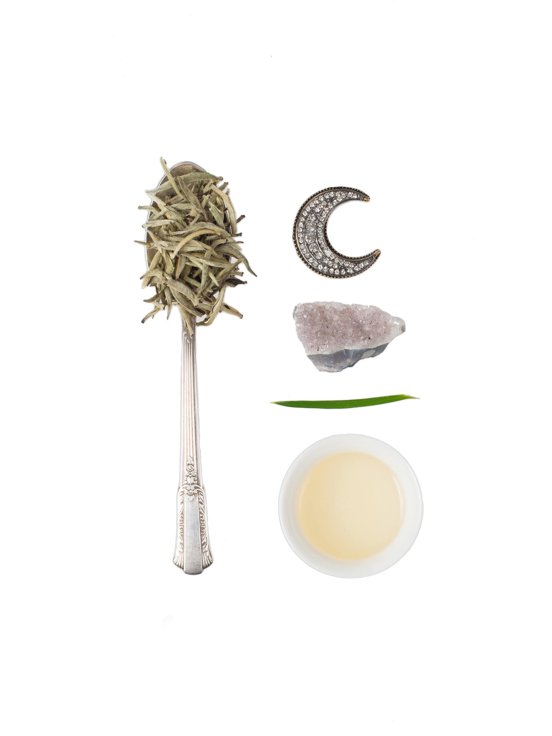 A flat-lay image shows a silver spoon filled with loose leaf tea, a crescent moon-shaped pendant, a raw amethyst crystal, a green leaf blade, and a small white bowl containing Magic Hour's Jasmine Moon White Tea of Reverence, all arranged on a white background.