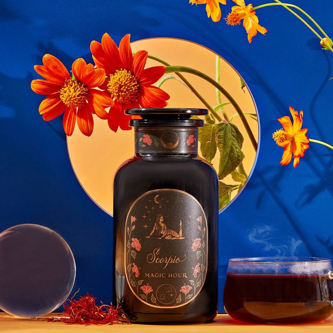 A dark jar labeled "Scorpio Tea for Sensual Brilliance" is placed on a table next to a teacup filled with Magic Hour Tea, saffron threads, and a clear orb. The background features bright orange flowers and a vibrant blue backdrop with a circular opening revealing more flowers.