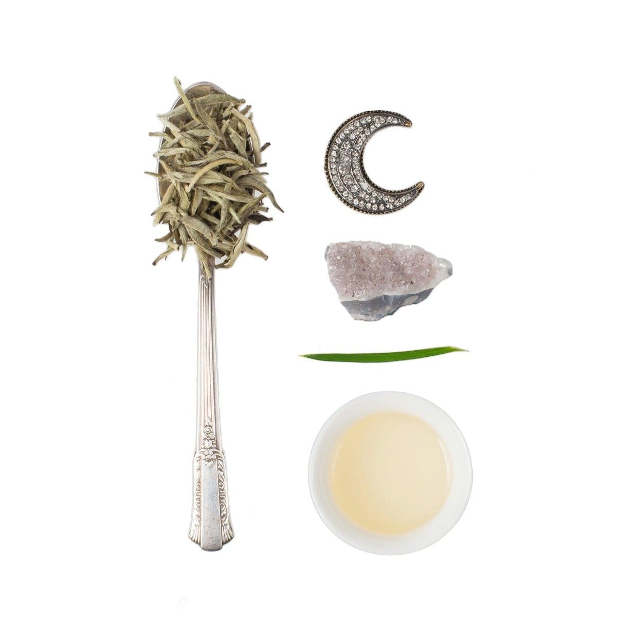 A flat lay image features a silver spoon filled with dried herbs, a beaded crescent moon, a small piece of amethyst crystal, a green leaf blade, and a white bowl containing light-colored Silver Moon White Tea from Club Magic Hour, all artfully arranged on a white background.