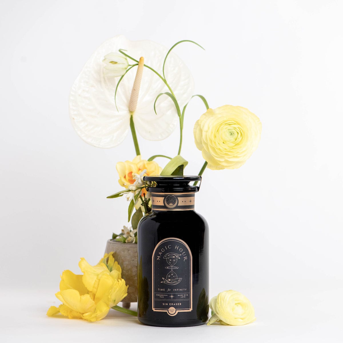 A black bottle labeled "Sin Eraser™ : Puerh Tea" by Magic Hour is surrounded by various flowers, including a white anthurium and yellow ranunculus, against a white background. The bottle has a gold-accented neck and the flowers visually frame it, adding an elegant touch.