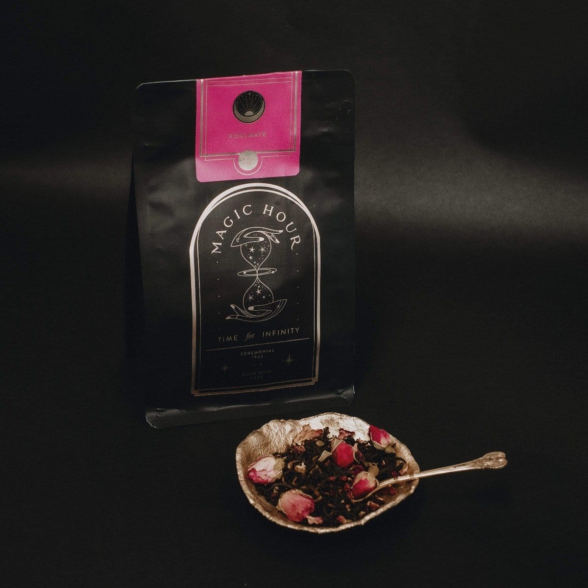 A package of Magic Hour Tea &quot;Soulmate: Chocolate-Raspberry-Rose Black Tea for Finding &amp; Celebrating Love&quot; with a pink label sits against a dark background. In front, a decorative bowl brims with organic tea leaves and dried rosebuds, while a small spoon rests on the bowl.