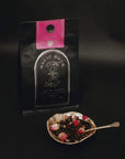 A package of Magic Hour Tea "Soulmate: Chocolate-Raspberry-Rose Black Tea for Finding & Celebrating Love" with a pink label sits against a dark background. In front, a decorative bowl brims with organic tea leaves and dried rosebuds, while a small spoon rests on the bowl.