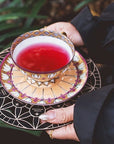 Person holding a Crystal Grid: Flower of Life Tea Ceremony Altar by Alibaba filled with organic red tea on a matching saucer, standing near lush green foliage, dressed in a dark coat.