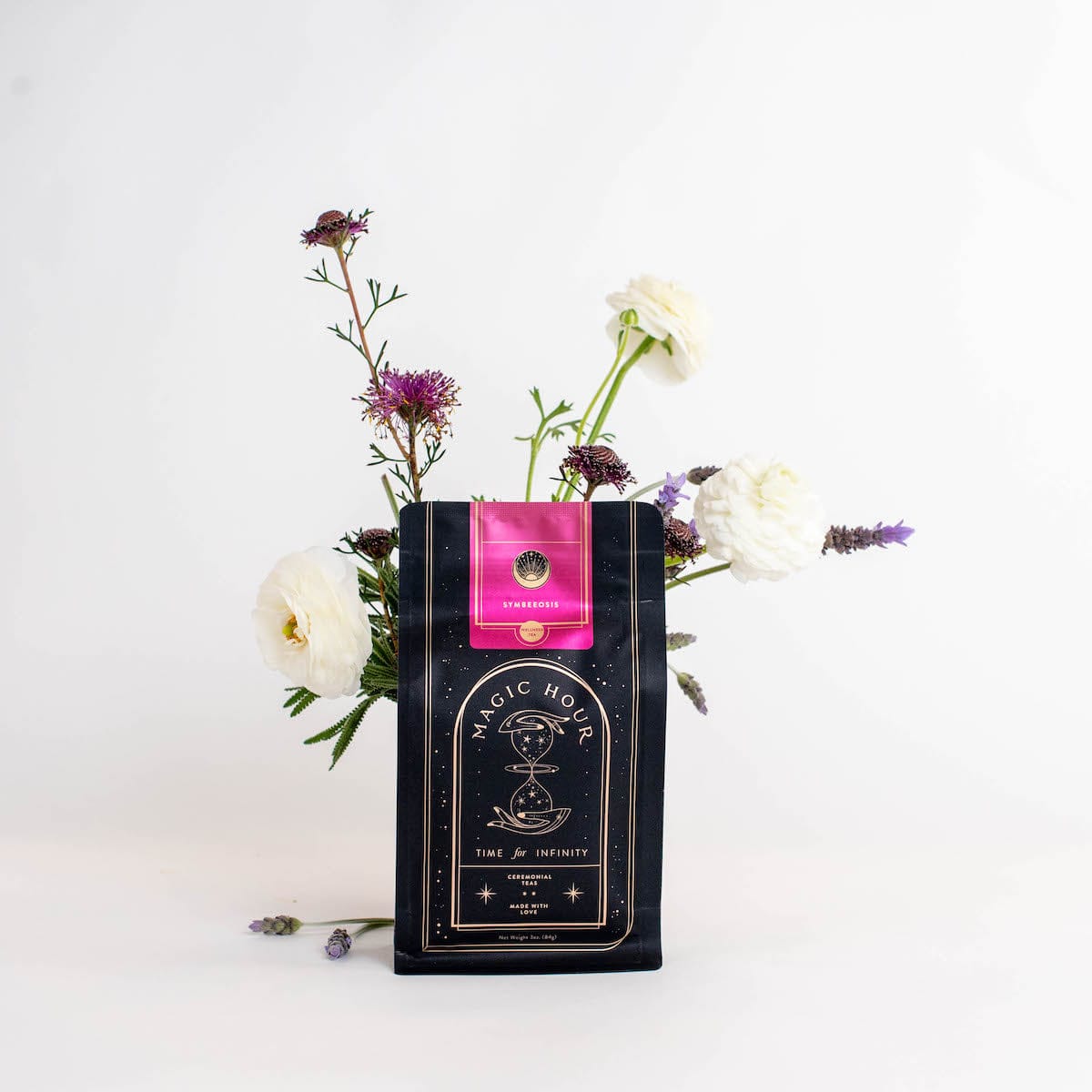 A black package of Magic Hour Symbeeosis: Beautifying Immunitea for the Queen Bee stands upright against a white background. The package is adorned with a pink label and surrounded by delicate flowers, including white ranunculus and purple blooms, arranged artfully to enhance the presentation.
