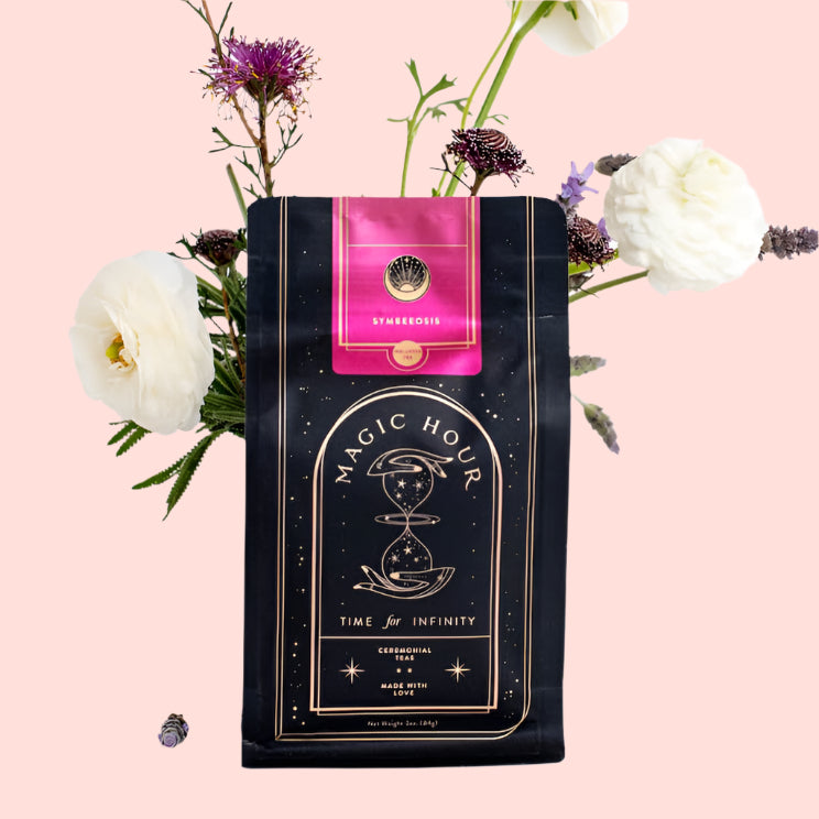 A black package of Magic Hour Symbeeosis: Beautifying Immunitea for the Queen Bee stands upright against a white background. The package is adorned with a pink label and surrounded by delicate flowers, including white ranunculus and purple blooms, arranged artfully to enhance the presentation.
