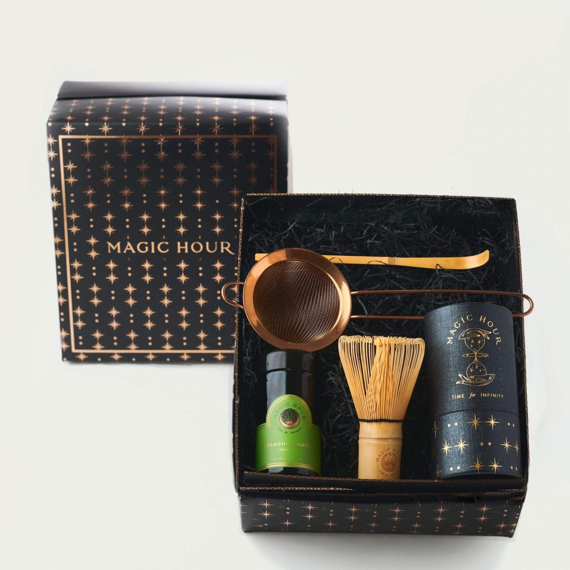 A gift box with a black and gold starry design featuring a bottle of organic tea, a matcha whisk, a gold tea strainer, and a box labeled "Ceremonial Matcha Traveler Gift Box" containing loose leaf tea. The box lid with the "Magic Hour" logo is partially removed and placed beside it.
