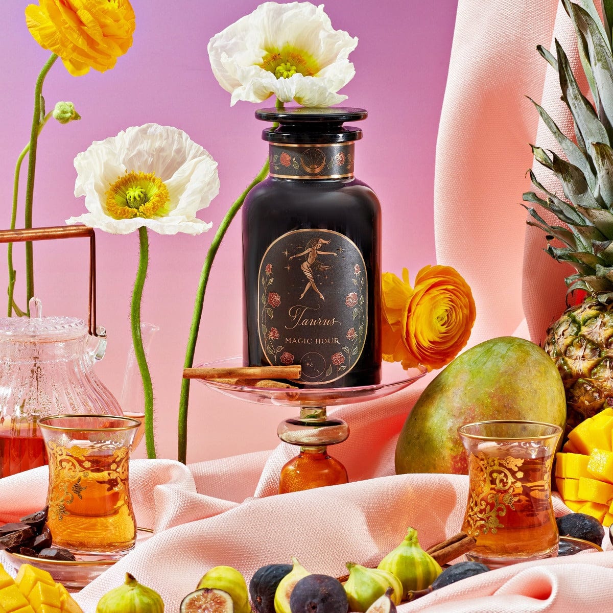 A decorative bottle labeled &quot;Taurus: Tea of Venusian Garden Delights&quot; from Magic Hour is surrounded by vibrant yellow and white flowers, fresh fruit including pineapple, mango, and figs, with a pink and purple gradient background. Two ornate glasses with amber liquid from the Magic Hour collection are placed in the foreground.