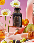 A decorative bottle labeled "Taurus: Tea of Venusian Garden Delights" from Magic Hour is surrounded by vibrant yellow and white flowers, fresh fruit including pineapple, mango, and figs, with a pink and purple gradient background. Two ornate glasses with amber liquid from the Magic Hour collection are placed in the foreground.
