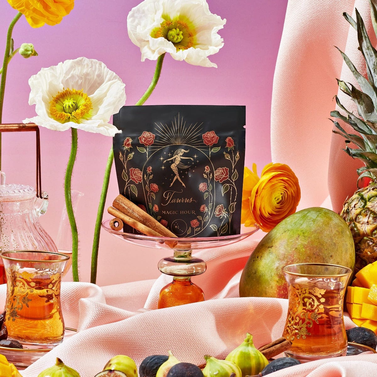 A vibrant still life featuring a black floral-embellished Magic Hour Taurus: Tea of Venusian Garden Delights package on a pink draped fabric, surrounded by tropical fruits like pineapples and mangoes, colorful flowers, two glass cups filled with organic tea, cinnamon sticks, and a sunset gradient background.