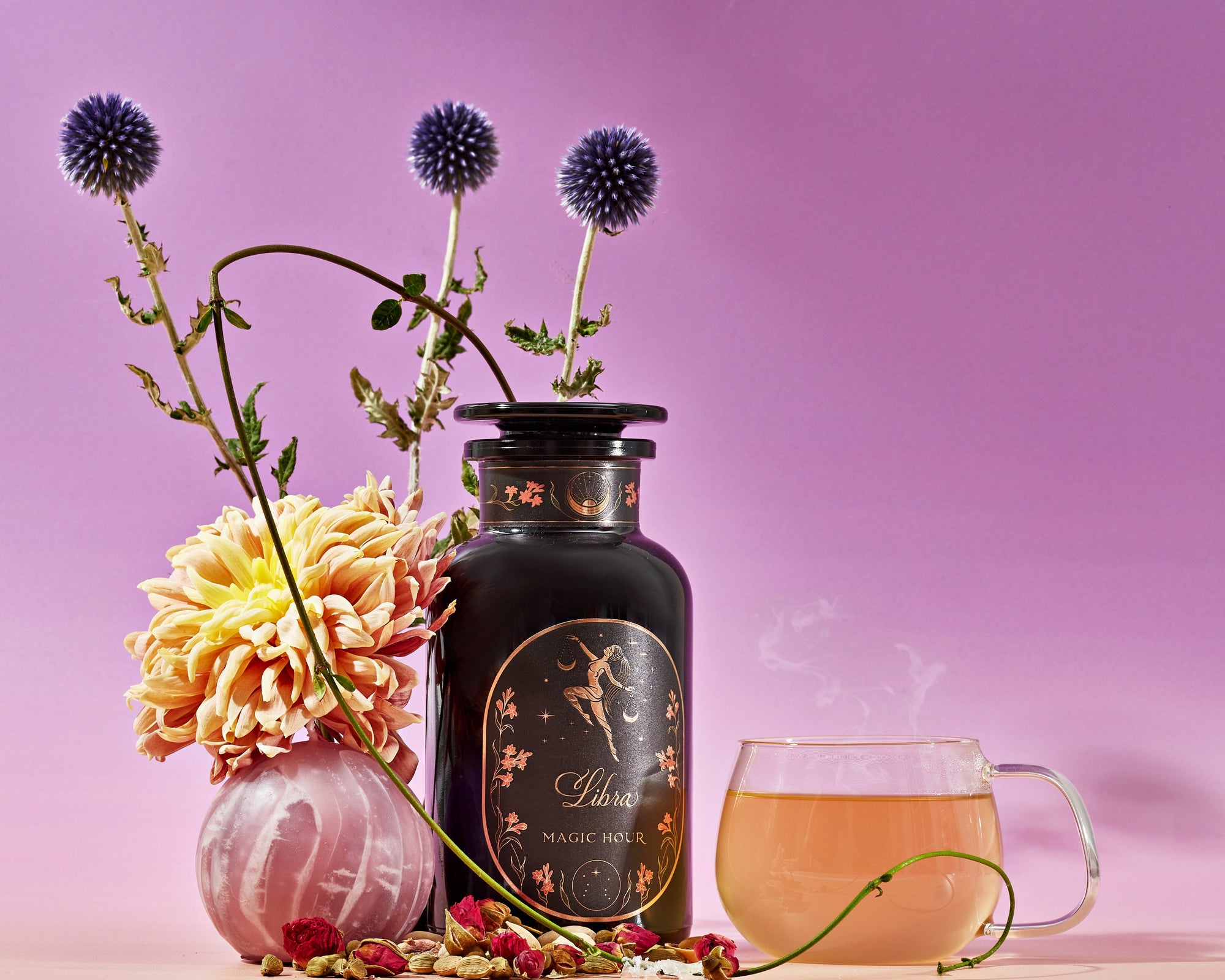 A pink and purple-toned still life image featuring a black bottle labeled "Magic Hour," adorned with intricate gold designs. Surrounding it are vibrant flowers, a glass teacup with steaming Libra- Pistachio-Rose Persian Love Cake Tea with White Pearls & Shatavari, dried herbs, and purple thistle-like flowers.