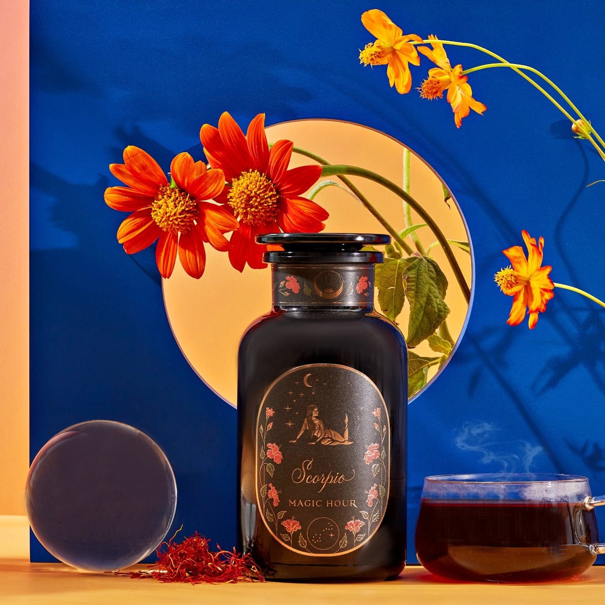 A dark jar labeled "Scorpio Tea for Sensual Brilliance" is placed on a table next to a teacup filled with Magic Hour Tea, saffron threads, and a clear orb. The background features bright orange flowers and a vibrant blue backdrop with a circular opening revealing more flowers.