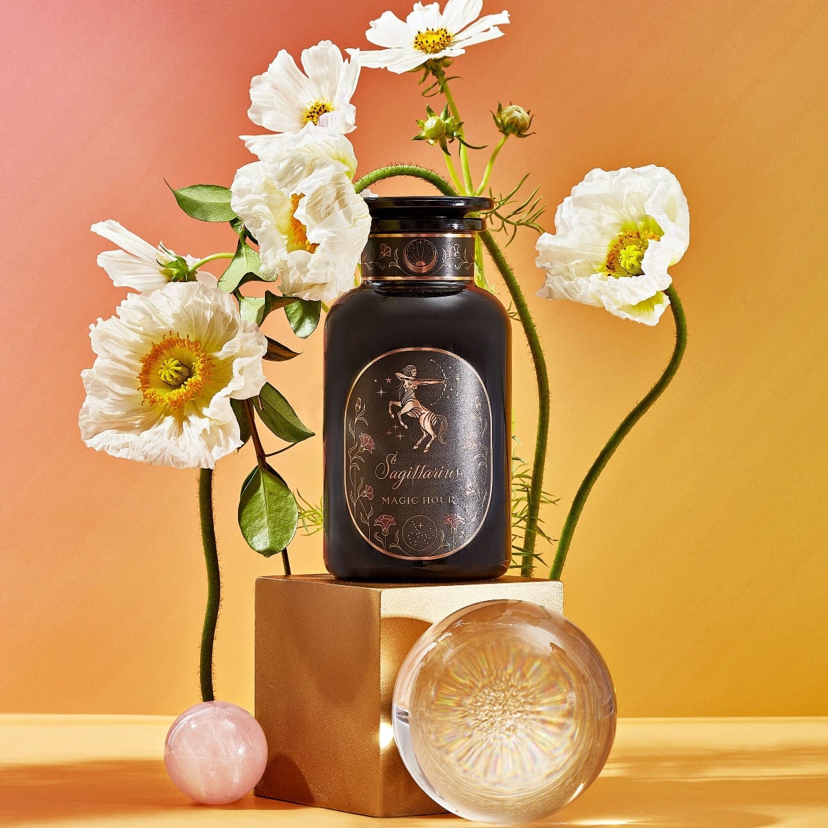 A black perfume bottle with a gold label reading "Sagittarius Tea of Good Fortune & Abundance - Magic Hour" is displayed on a gold pedestal against a gradient background of orange and pink. White flowers and two clear spheres, one pink and one iridescent, surround the bottle, evoking the elegance of Magic Hour tea rituals.