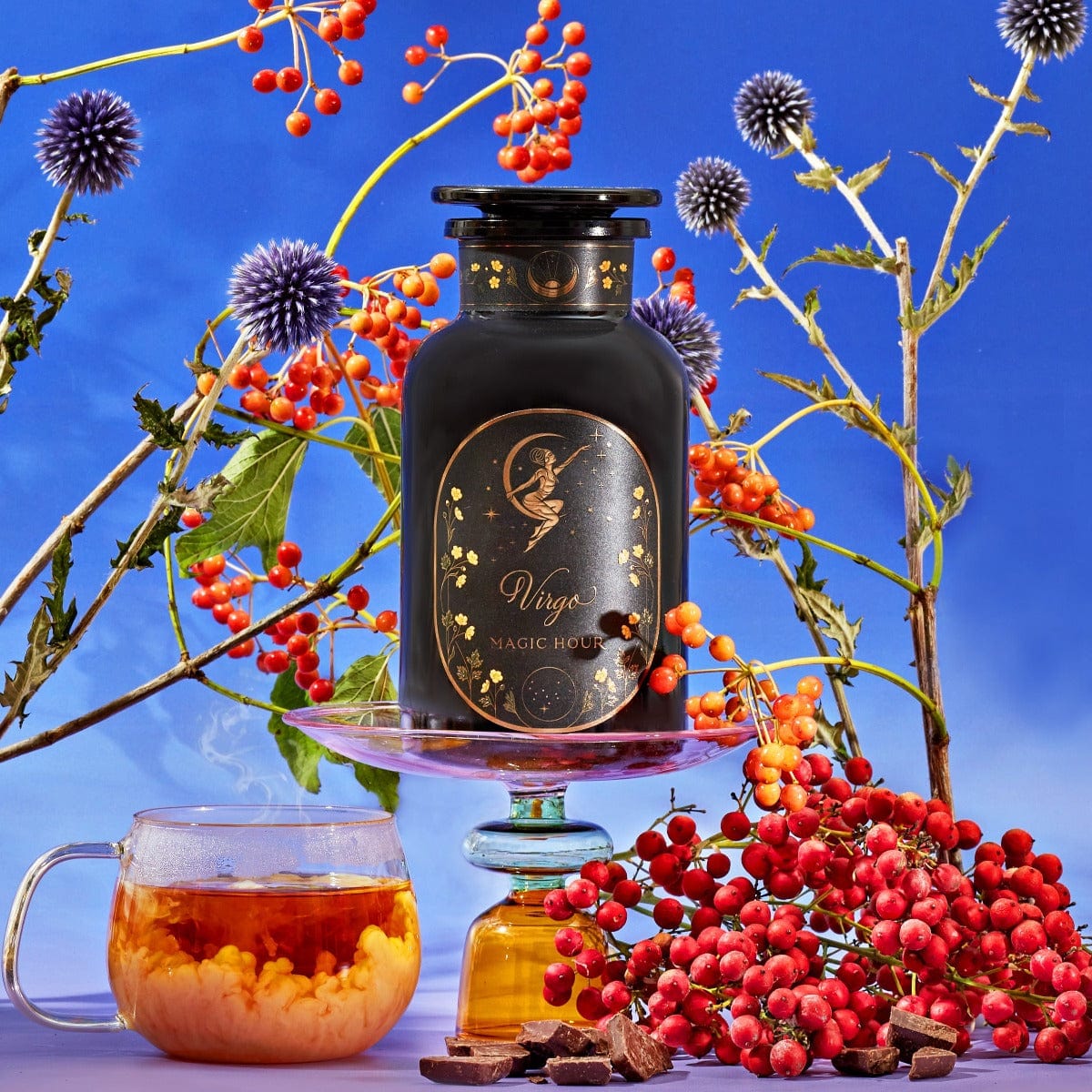 A black jar labeled "Magic Hour Virgo Tea of Virtue, Wit & Meticulous Magic" sits on a glass stand among vibrant orange berries, purple thistle flowers, and leafy branches under a blue sky. Beside it, a clear glass mug of Organic Tea with loose leaf tea and a cluster of red berries complete the harmonious arrangement.